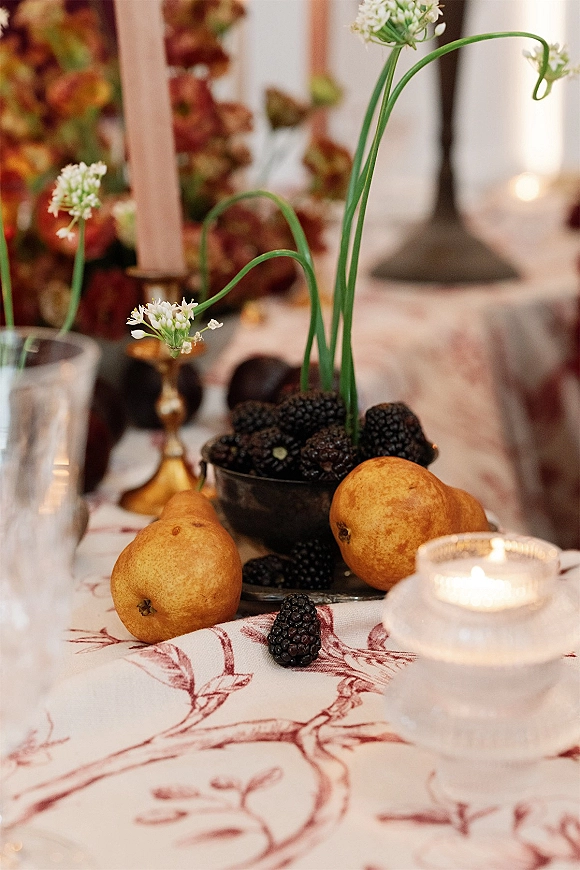 Reception tablescape with fruit wedding centerpiece featuring pears, blackberries, taper candles, and white blooms on a patterned cloth in warm light