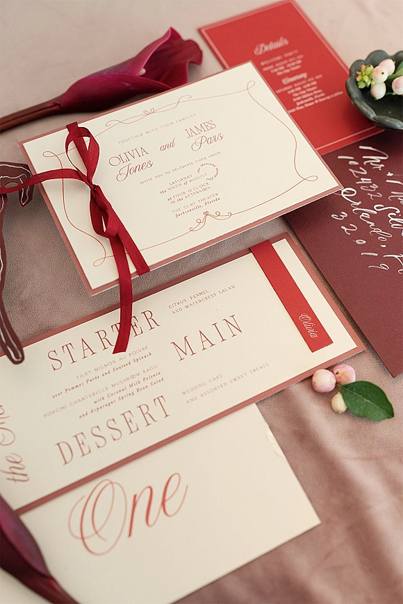 Invitation suite with burgundy wedding invitations tied with a burgundy ribbon, wax seal, calla lily and greenery on linen backdrop