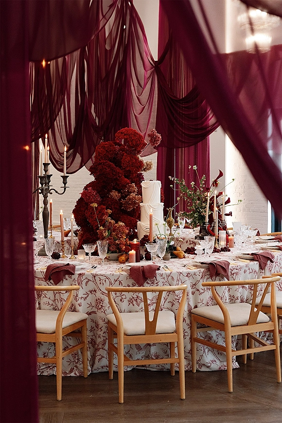Reception tablescape with burgundy wedding tablescape draping, taper candles, floral centerpiece with grapes and pears, and a white tiered cake by window light