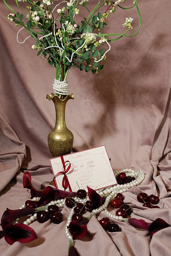 Invitation suite wedding invitation flat lay with red ribbon and pearl accents, styled with calla lilies and cherries on draped mauve fabric