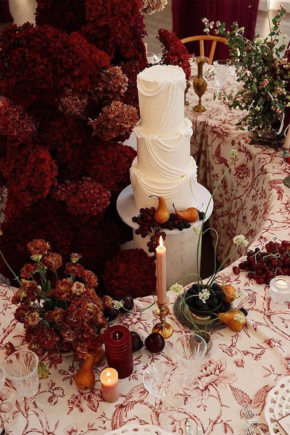 Wedding cake with draped buttercream on a stand, styled with grapes and pears, surrounded by brass candlesticks and red florals at the reception table