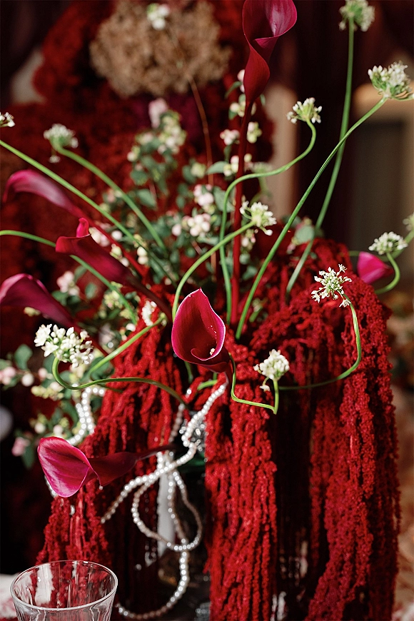 Wedding centerpiece with red calla lily centerpiece blooms in burgundy and white, greenery, and pearl strands spilling from a tall glass vase indoors