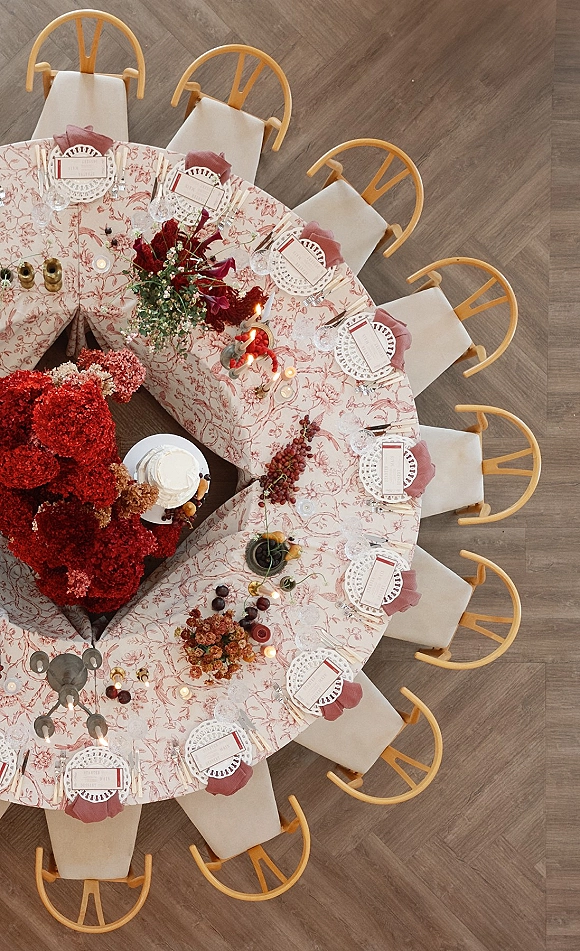 Reception tablescape with a round wedding table setup on patterned tablecloth, candles, red floral and grape centerpieces, wood floor beneath