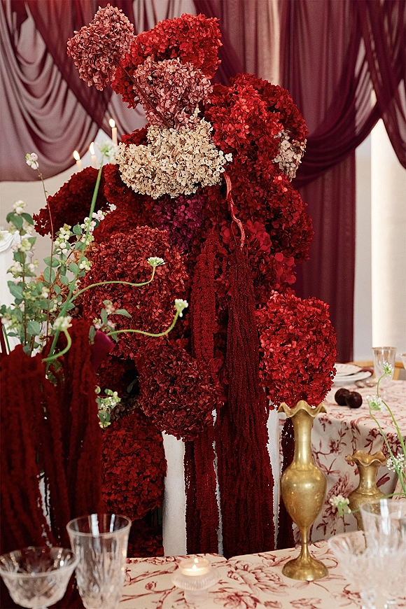 Reception centerpiece with red hydrangea blooms and hanging amaranthus in gold vases, surrounded by taper candles on a patterned tablecloth.