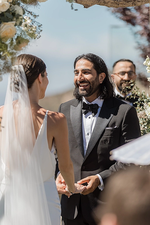 Wedding vows as bride and groom hold hands under a floral arch, her long veil and sleek gown facing his black tuxedo in blue-sky landscape