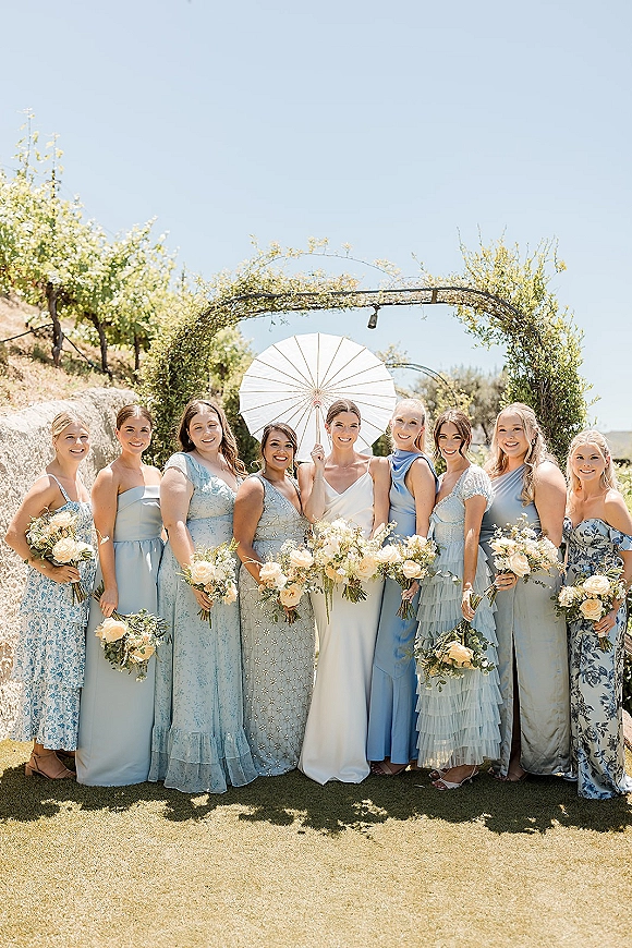 Bridesmaid group photo with bride and bridesmaids in blue dresses holding white and blush bouquets under a greenery arch with string lights at a vineyard lawn