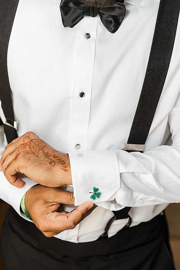 Groom getting ready adjusts a green shamrock cufflink on a white tuxedo shirt, black bow tie and suspenders visible indoors