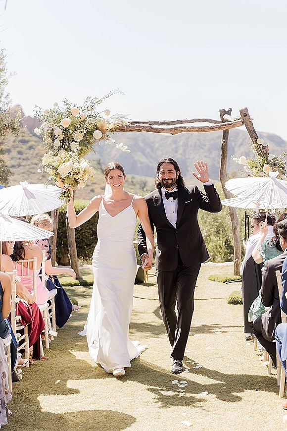 Wedding recessional as bride and groom walking down aisle, holding hands and waving under a floral arch with mountain view and parasols