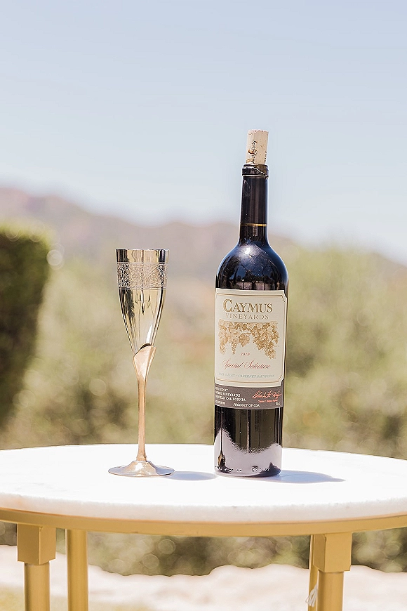 Wedding wine detail with a red wine bottle, champagne flute, and cork on a small table against blurred hills and sky outdoors