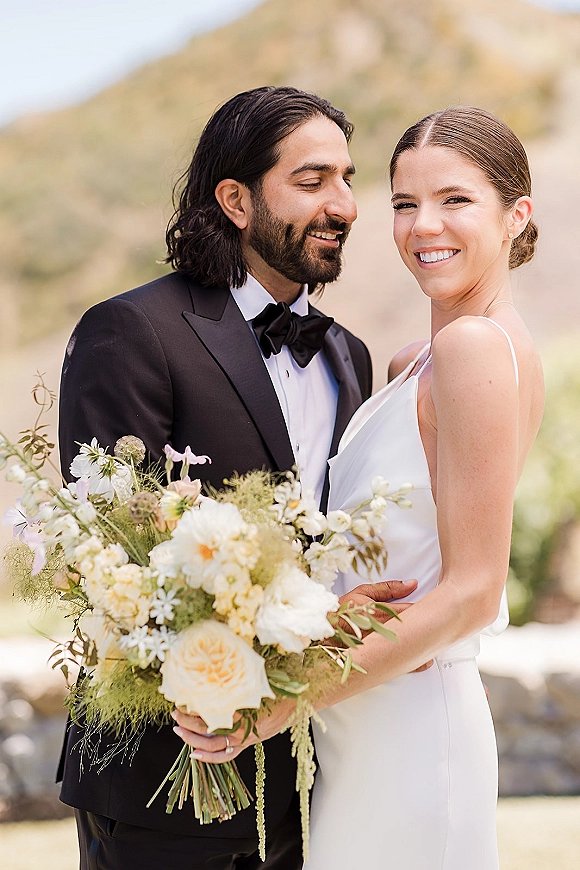 Couple portrait of bride and groom portrait, she holds a bridal bouquet in a satin dress as he wears a tuxedo with mountain hills behind