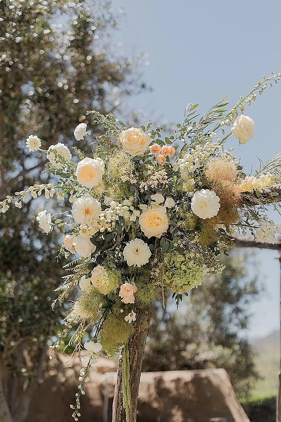Wedding altar flowers with ceremony arbor blooms of white and peach roses, hydrangea, and eucalyptus on a wooden post under blue sky