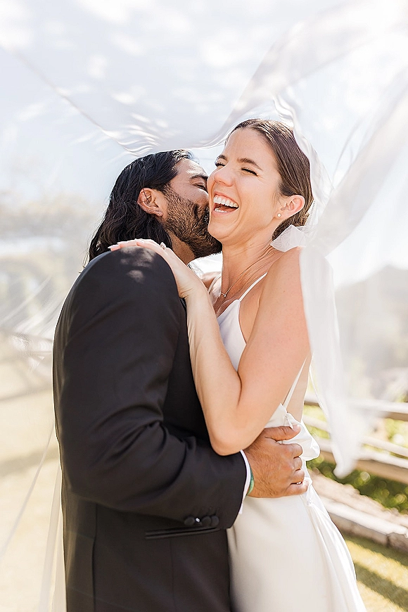 Wedding couple portrait with groom kissing bride’s cheek as she laughs, bridal veil blowing in the wind by mountains and a wooden fence