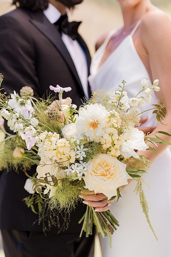 Wedding bouquet with white and peach bridal bouquet blooms and greenery held by bride, with groom in black tuxedo against blurred greenery
