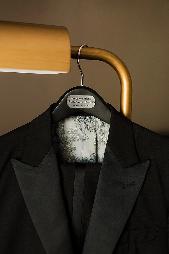 Groom suit detail with a black tuxedo jacket on a custom engraved hanger, patterned lining visible on a brass rack against a neutral wall