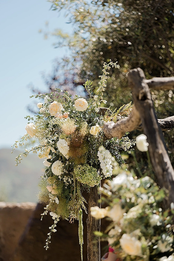 Ceremony floral arrangement with white roses and hydrangea draped on a rustic wooden fence, greenery and amaranthus in sunlit trees