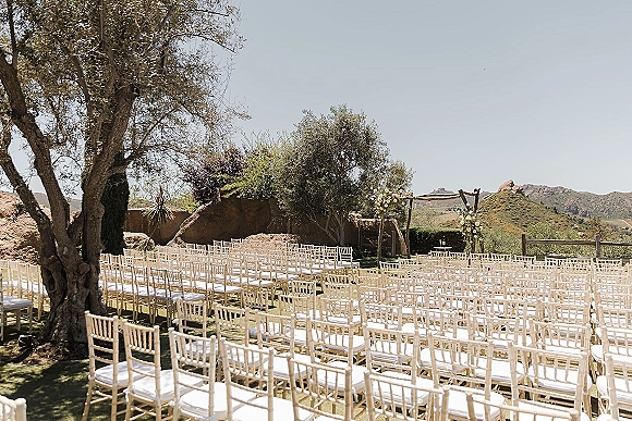 Ceremony setup with outdoor ceremony seating of white Chiavari chairs facing a wooden altar arch with florals on a lawn beneath mountains and olive trees