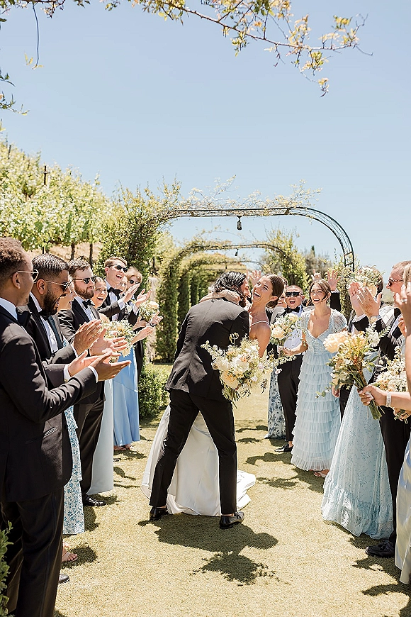 Wedding recessional as newlyweds walk the aisle under a floral arch tunnel, wedding party in sunglasses cheering on a sunny garden lawn