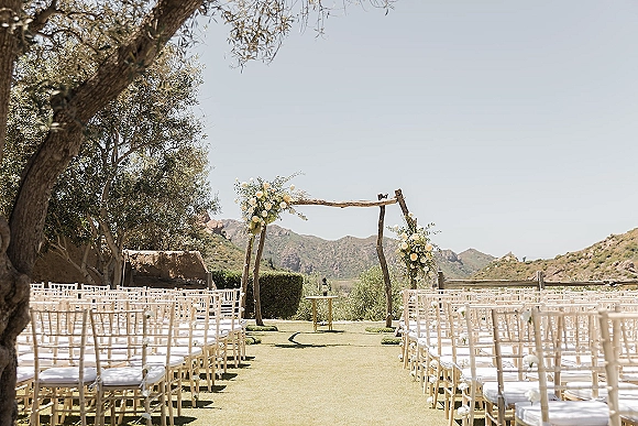 Ceremony setup with an outdoor ceremony aisle leading to a wood wedding arch with white roses and greenery, set on a lawn with mountain views
