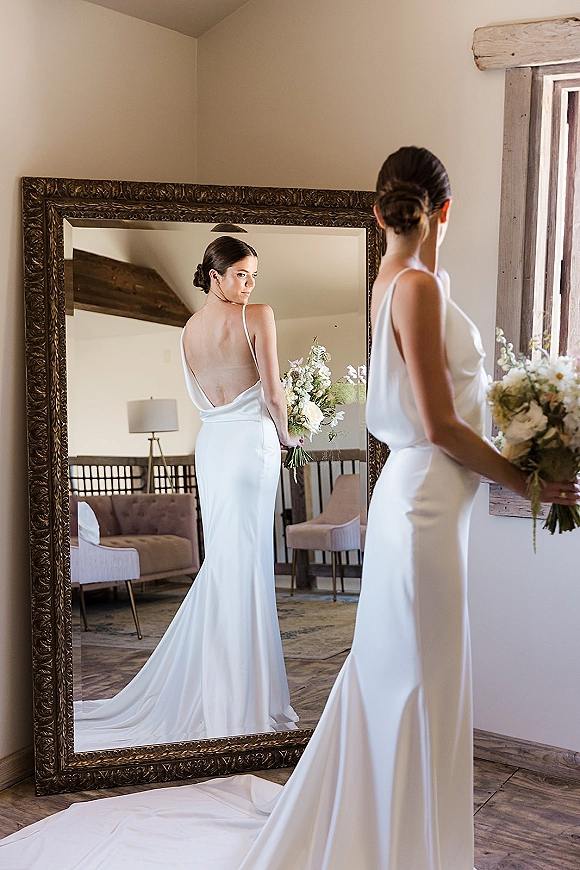 Bridal portrait of a bride in mirror holding a white bouquet in a low back wedding dress, standing by a window in a cozy suite