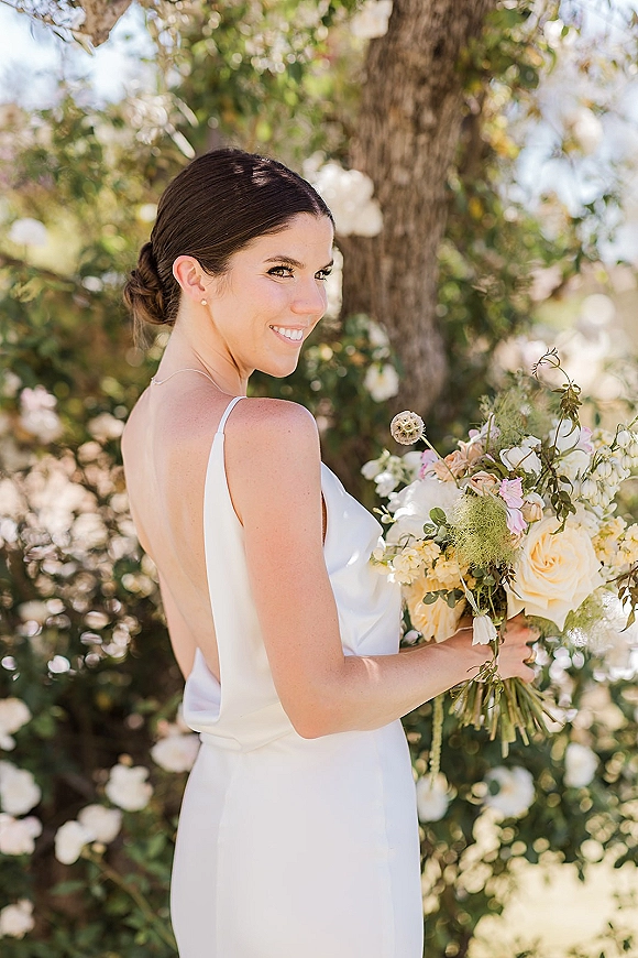 Bridal portrait of a bride holding bouquet of roses and greenery, looking over her shoulder in sunlight beneath a flowering tree