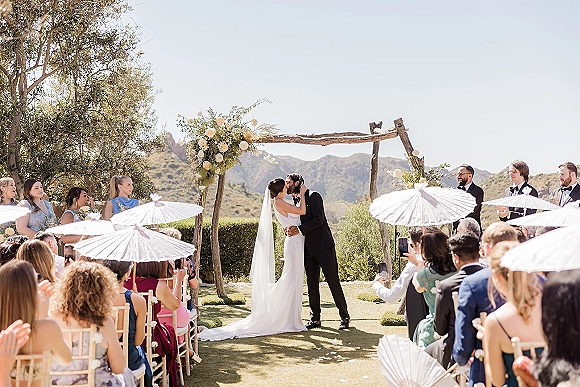 Ceremony kiss as bride and groom embrace under a floral arch, veil flowing, guests with paper parasols, mountains behind