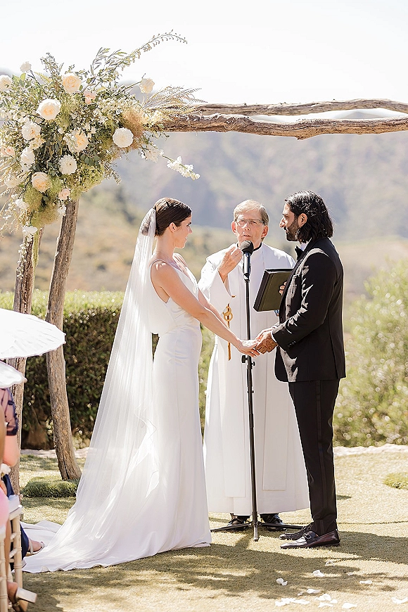 Wedding vows as bride and groom hold hands beneath a floral arch, officiant reading into microphone with mountain view behind