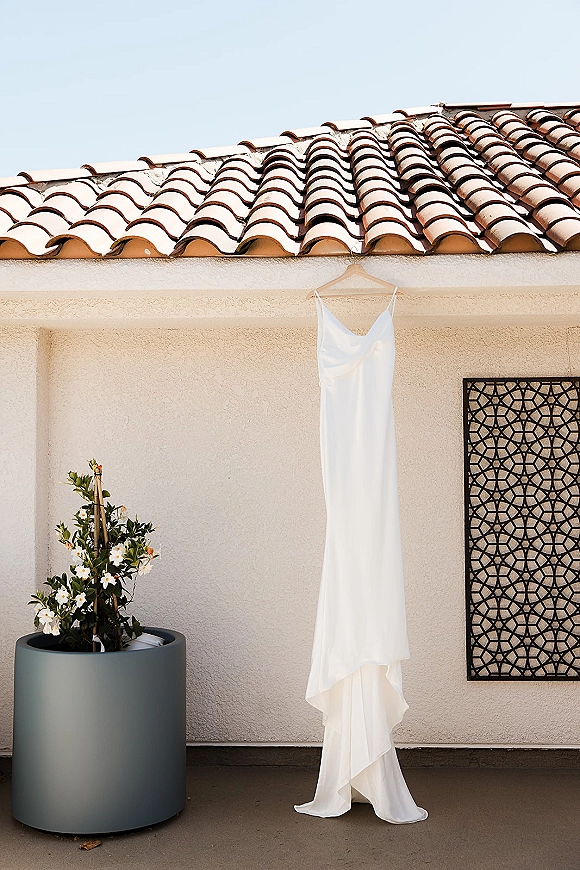 Wedding dress on hanger with a potted flowering plant accent against a stucco wall patio, with clay tile roof and sky above