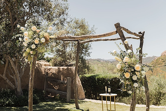 Wedding ceremony arch with a rustic wooden wedding arch, asymmetrical white and blush roses and hanging greenery on a lawn with hills beyond