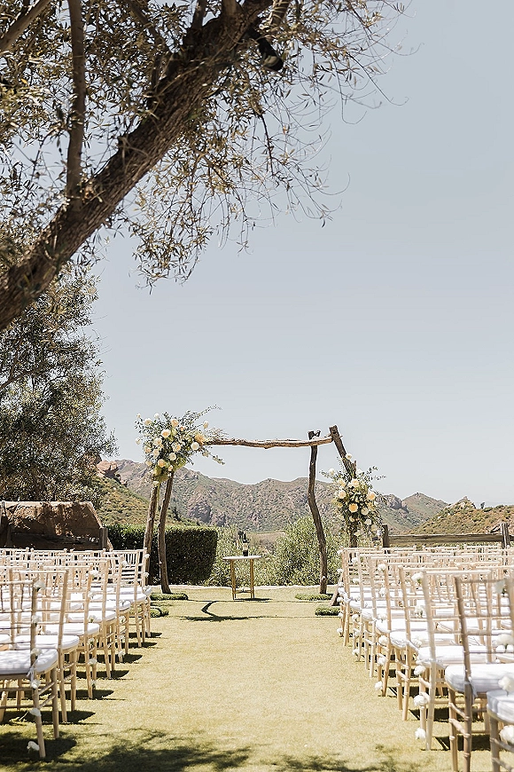 Ceremony setup for an outdoor wedding ceremony with a wood wedding arch, white roses and greenery on a grass aisle with mountain views