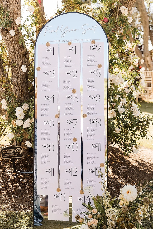 Wedding seating chart arched find your seat sign with calligraphy escort cards and wax seals on an easel, accented by florals in a sunlit garden