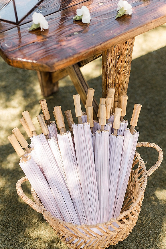 Wedding parasols in a woven basket on a rustic wooden table with white petals and a glass terrarium on a grassy lawn
