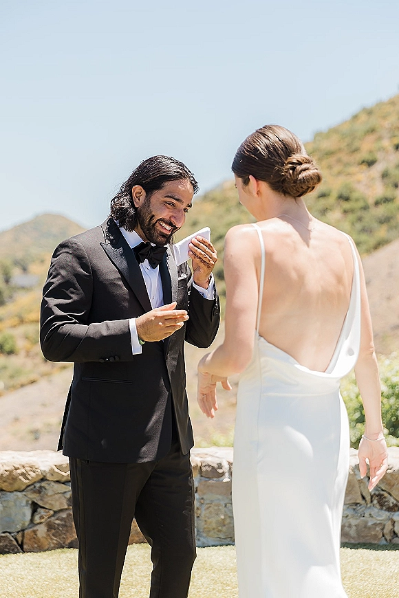 First look moment as bride in an open-back gown approaches groom in black tuxedo wiping tears beside a stone wall with mountain hills behind