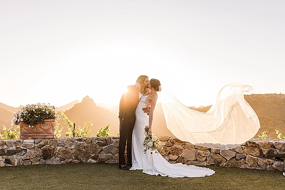 Wedding kiss portrait of the bride and groom kissing at sunset, her veil blowing in the wind as mountains glow behind a stone wall