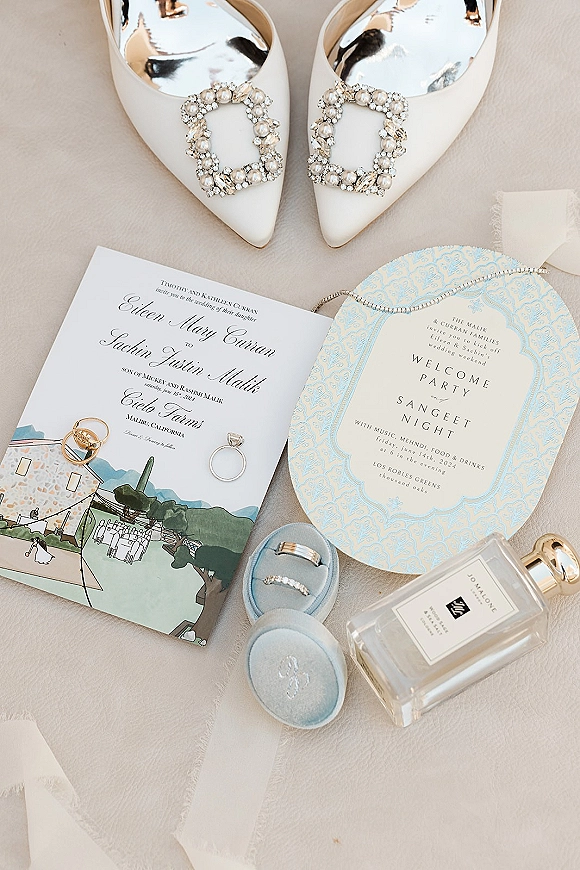 Invitation suite wedding flat lay with blue stationery, crystal buckle bridal shoes, rings and perfume arranged on a neutral fabric backdrop