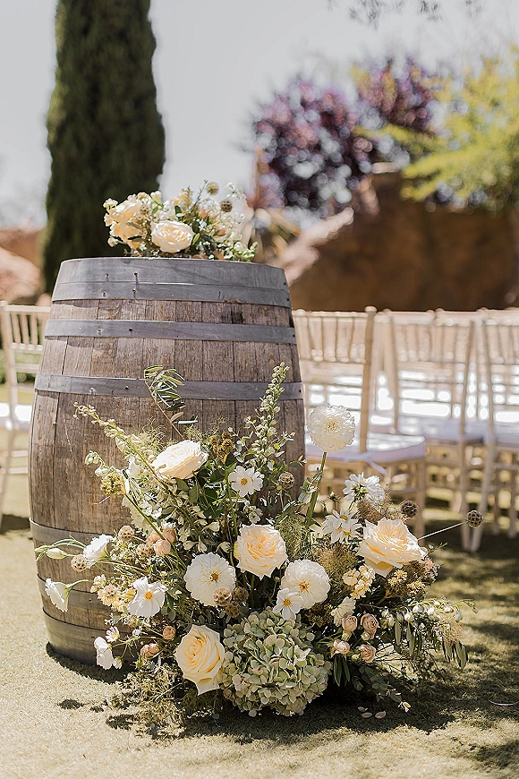 Ceremony aisle decor with a wine barrel wedding decor floral arrangement of cream roses, hydrangea, and petals on an outdoor lawn aisle