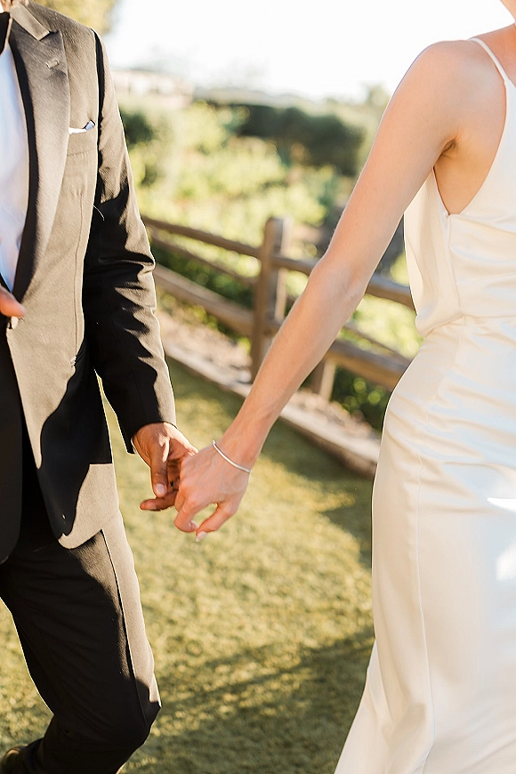 Couple holding hands as bride and groom walking away across an outdoor lawn, wedding dress and black suit beside a rustic fence and hills