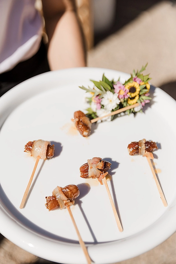 Wedding cocktail hour appetizers, passed hors d'oeuvres wedding on wooden skewers with bacon-wrapped bites and edible flower garnish on a white plate outdoors