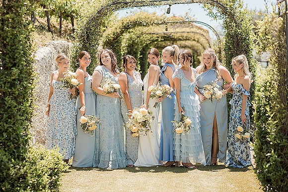 Bridesmaid group photo with blue bridesmaid dresses, holding blush and white rose bouquets under a vine-covered archway with string lights