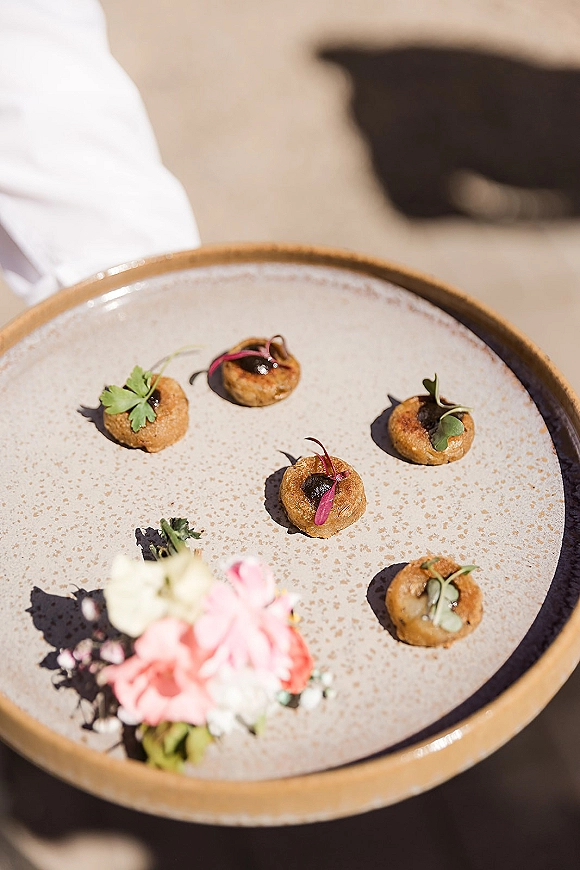 Wedding appetizers arranged as passed hors d'oeuvres on a ceramic platter, bite-size canapés topped with microgreens in sunlit outdoors with guests blurred