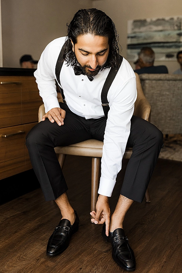 Groom getting ready, sitting to put on black loafers in a hotel room, wearing a white shirt with black bow tie and suspenders