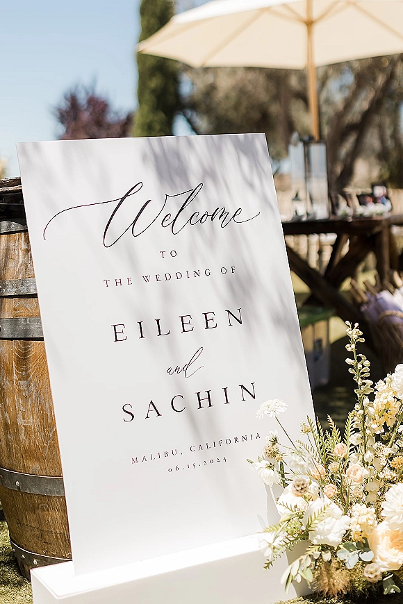 Wedding welcome sign acrylic with modern calligraphy on a wooden barrel, topped with blush and cream flowers on an outdoor patio under blue sky
