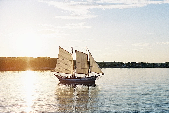Sailboat at sunset gliding on calm water with sails and rigging silhouetted, sun glare on the horizon near distant hills
