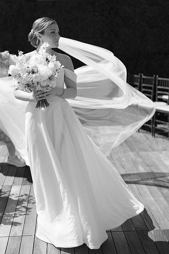 Bridal portrait in black and white of a bride holding bouquet, veil blowing as she looks away on an outdoor wooden deck by greenery