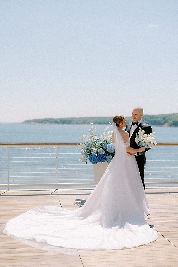 Couple portrait at a waterfront wedding, bride in a long-train dress and veil embracing groom in tuxedo on an ocean-view terrace deck