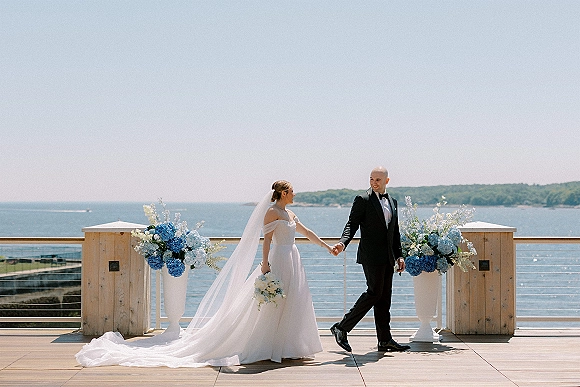 Couple portrait of bride and groom holding hands on a wooden deck, her long veil trailing beside blue hydrangea florals with ocean backdrop