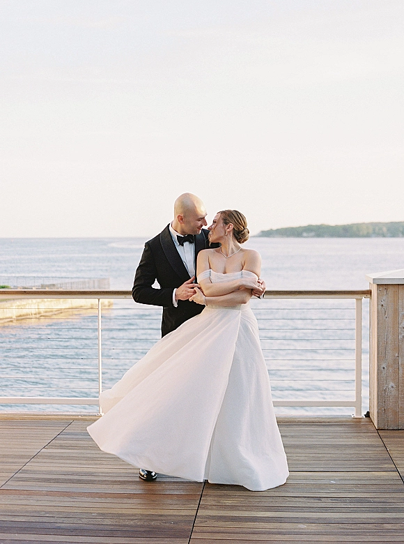 Couple portrait of bride and groom embrace on a wooden deck, her off-shoulder gown and his black tuxedo framed by ocean horizon