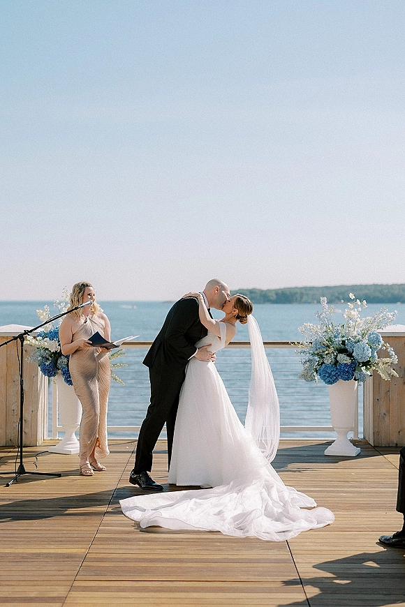 Ceremony kiss as bride in cathedral veil and strapless dress kisses groom in tux on oceanfront deck with hydrangea urns