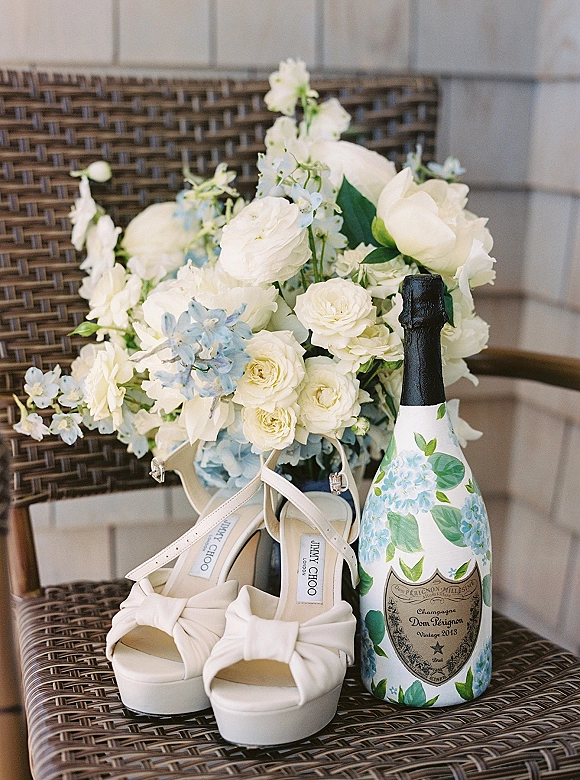 Bridal details flatlay featuring wedding detail styling with ivory bow heels, a white and blue bouquet, and champagne by a wicker chair