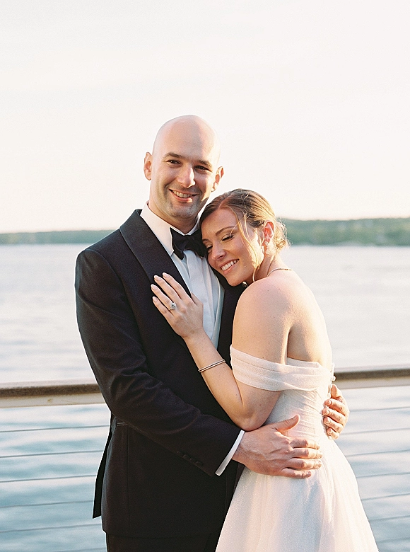 Couple portrait of bride and groom hug, her head resting on his shoulder, by a calm lakeshore with sky and distant trees, engagement ring visible