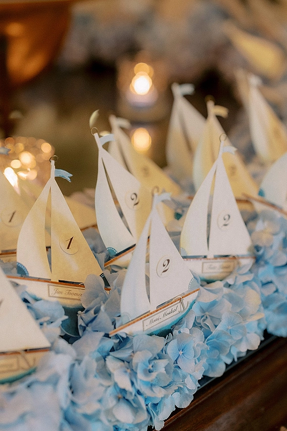 Wedding escort cards folded into paper sailboats with table numbers on a wooden tray, accented by blue hydrangeas and candlelight